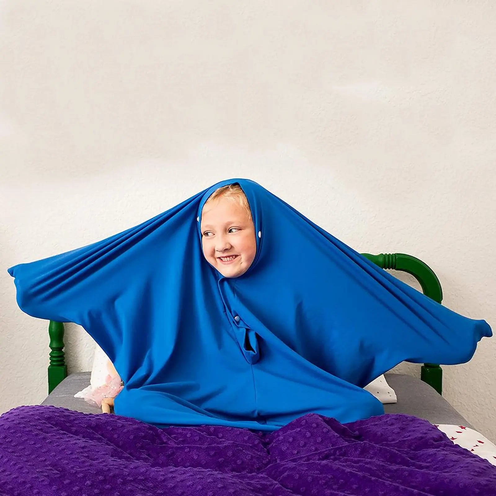 Child wearing a blue hooded garment sitting on a bed with a purple blanket.